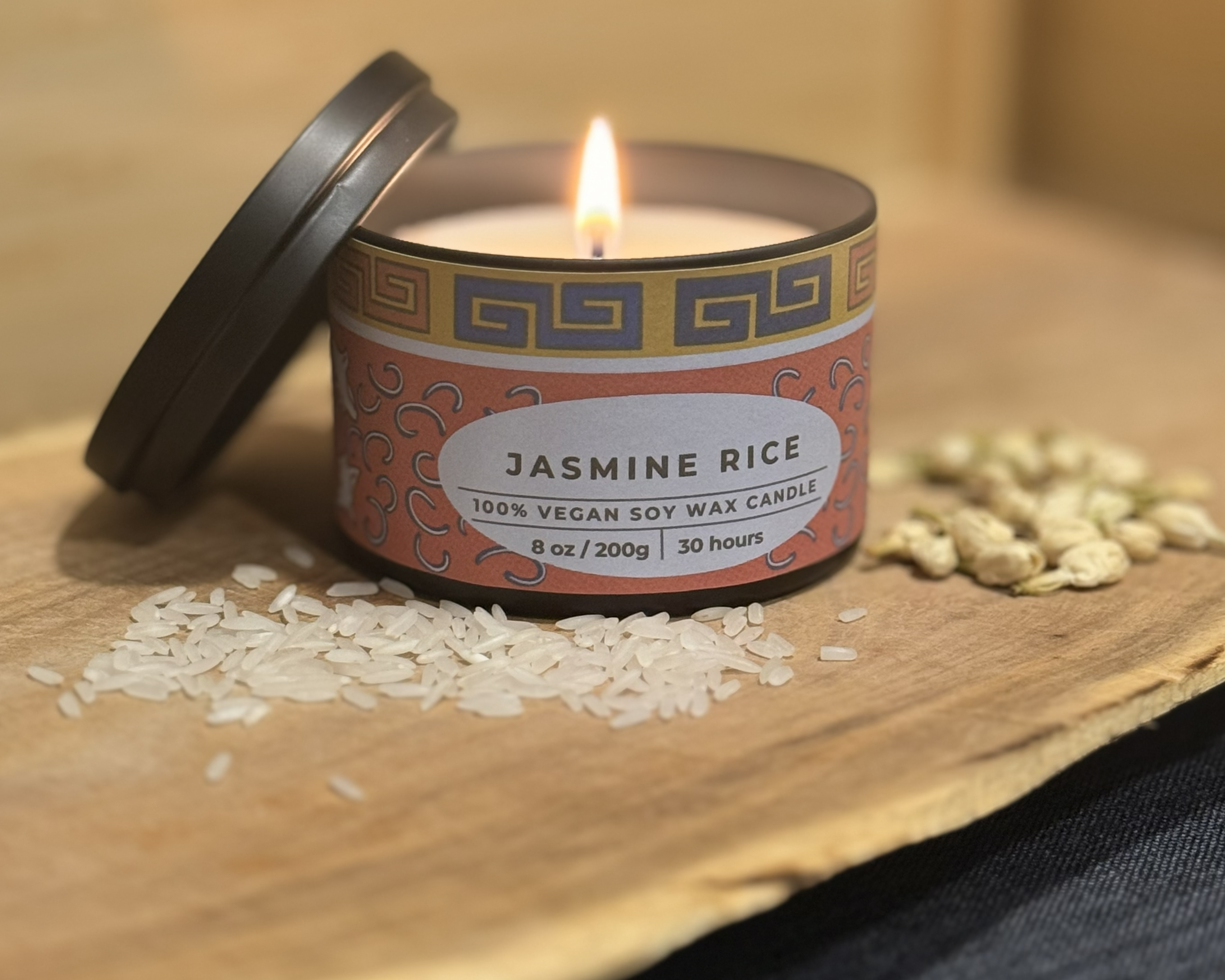 Candle labeled 'Jasmine Rice' with a lit wick on a wooden surface with rice grains.